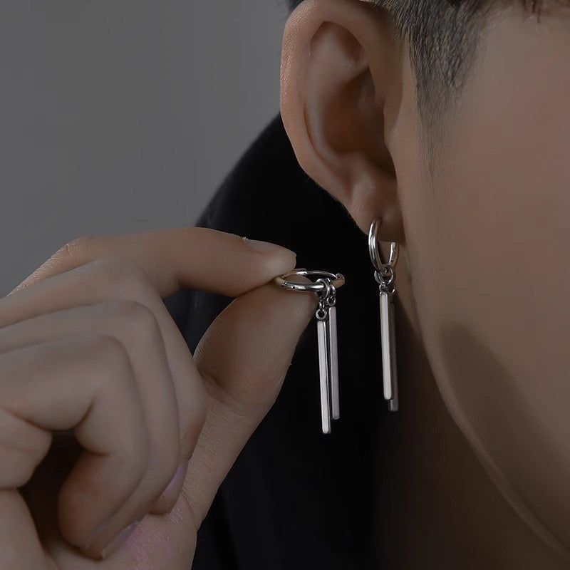 Person wearing a silver earring with dangling bars, held by a hand against a dark background.